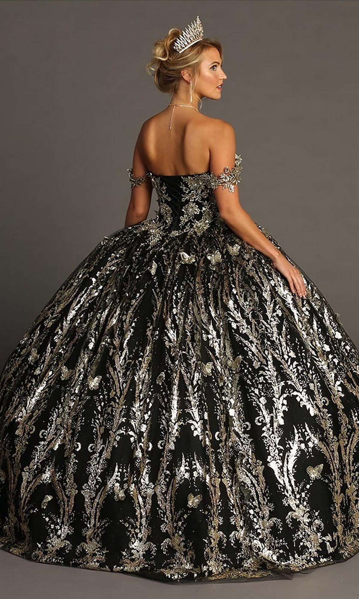 A woman stands gracefully against a plain background, wearing the Quinceanera Dress CHF3101 By Chicas—an ornate black and silver off-the-shoulder gown featuring a dramatic tulle ballgown skirt and delicate metallic-silver embellishments. Her look is completed by a sparkling tiara.